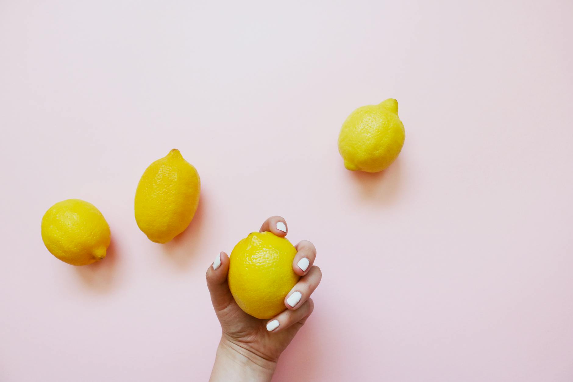 Une main tenant un citron jaune vif sur un fond rose pastel, entourée de trois autres citrons frais